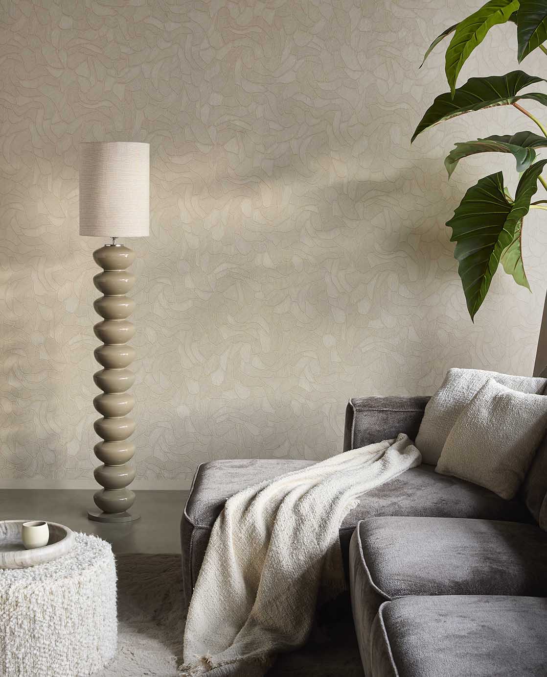 Pebble Relief wallpaper in beige behind a grey sofa, floor lamp and plant in a living room