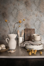 Rost wallpaper by Sandberg in grey patina styled behind a kitchen bench with a white jug, stacked ceramics and dried billy buttons