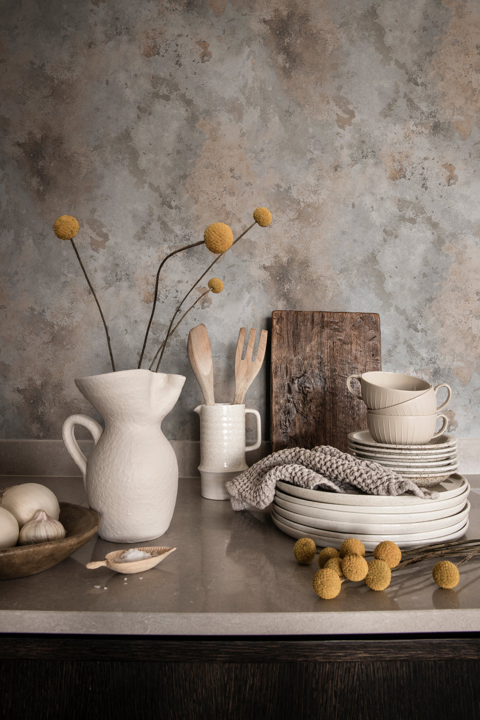 Rost wallpaper by Sandberg in grey patina styled behind a kitchen bench with a white jug, stacked ceramics and dried billy buttons