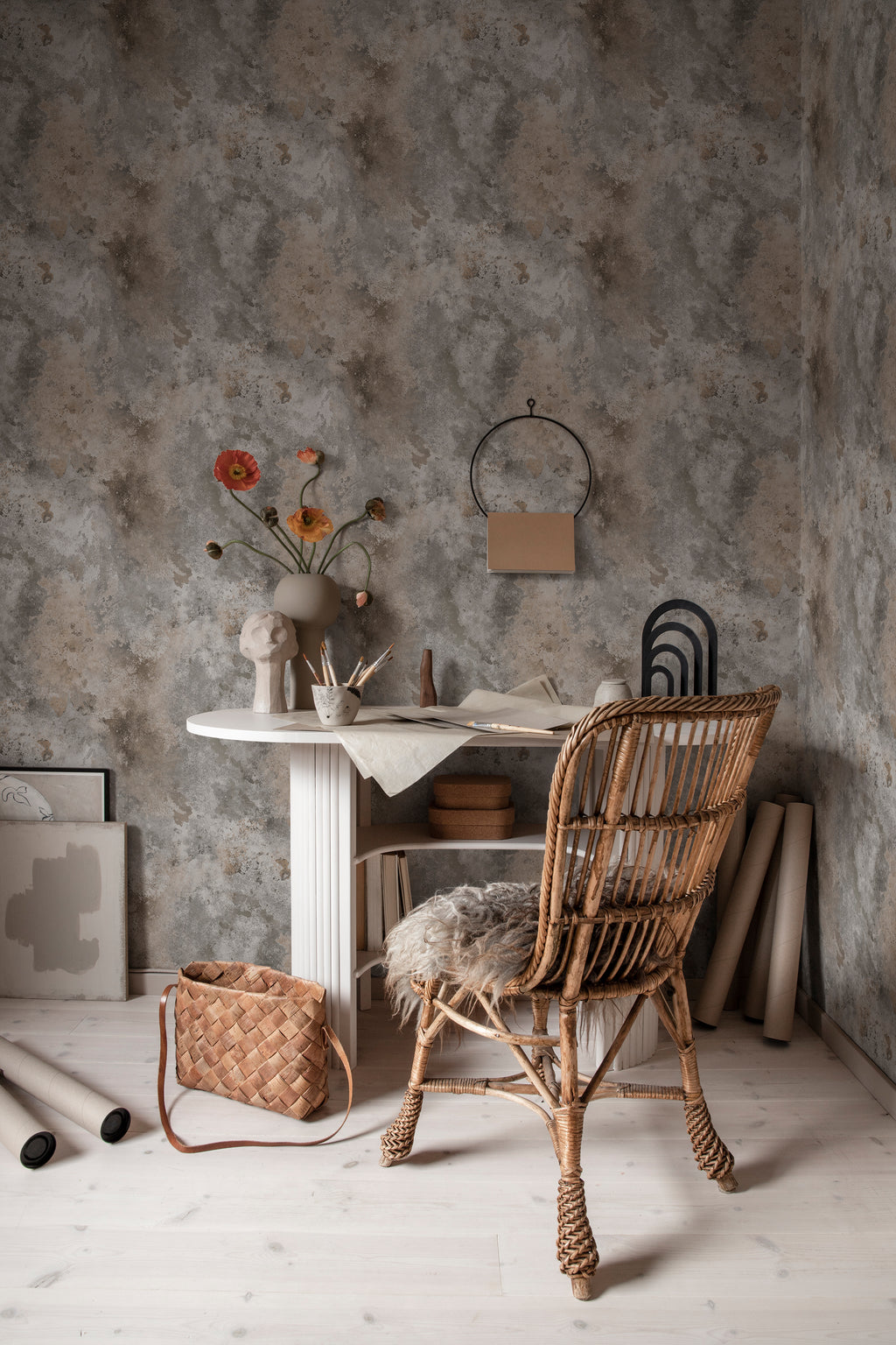 Rost wallpaper by Sandberg in mottled grey styled in a home office with a white desk, rattan chair and rolled paper