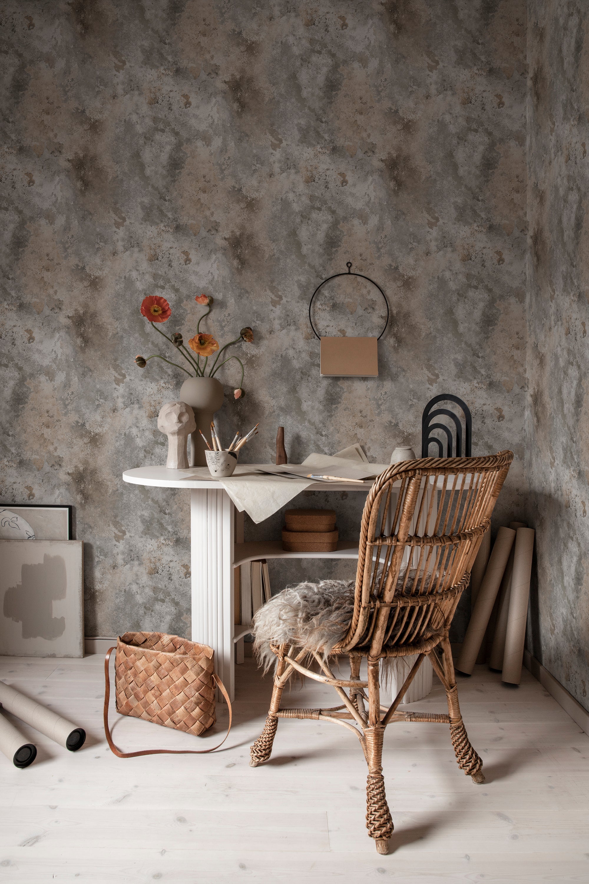 Rost wallpaper by Sandberg in mottled grey styled in a home office with a white desk, rattan chair and rolled paper
