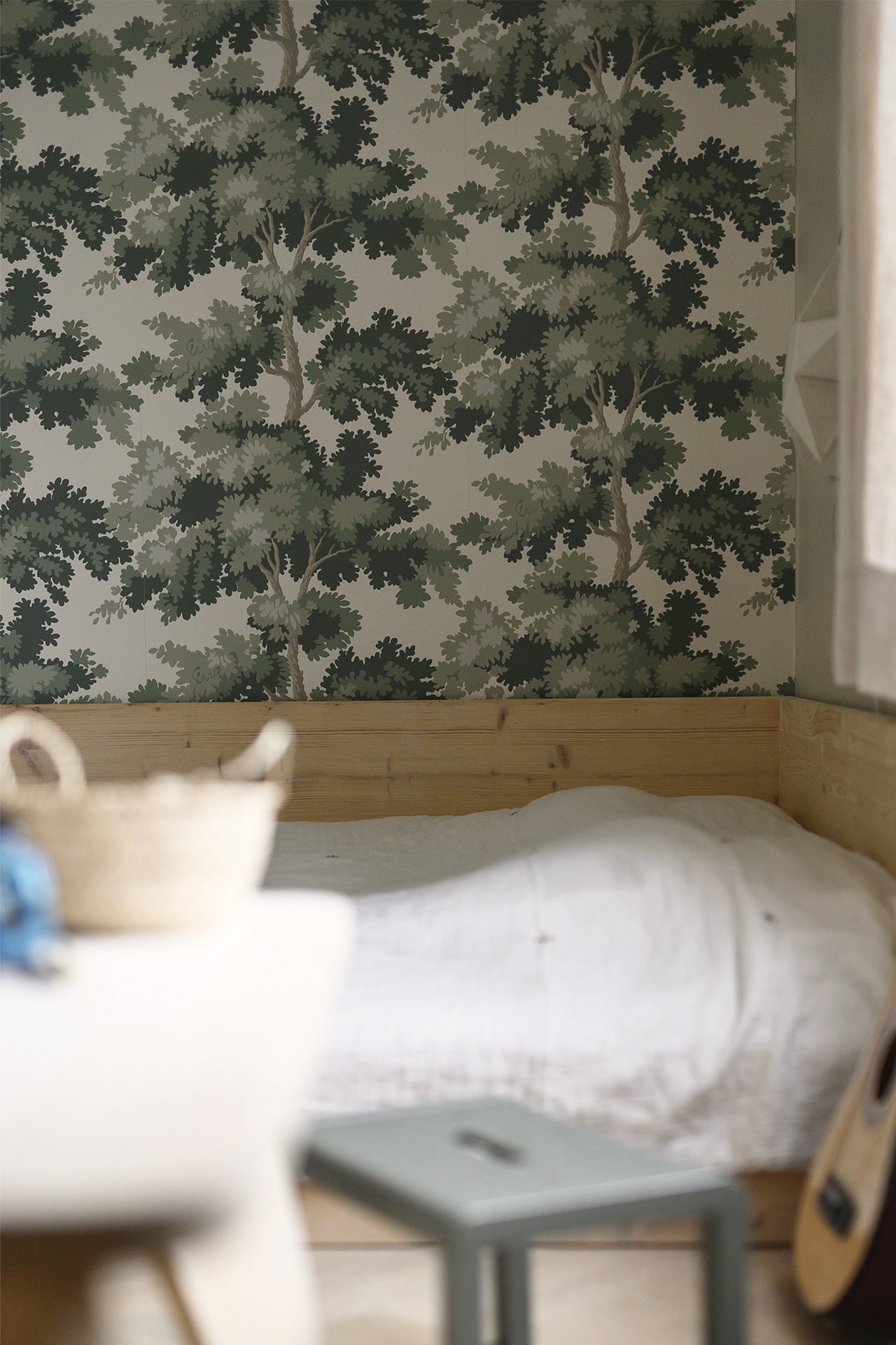 Raphael wallpaper by Sandberg in Green in a bedroom corner with white bed linen, timber paneling and a woven basket