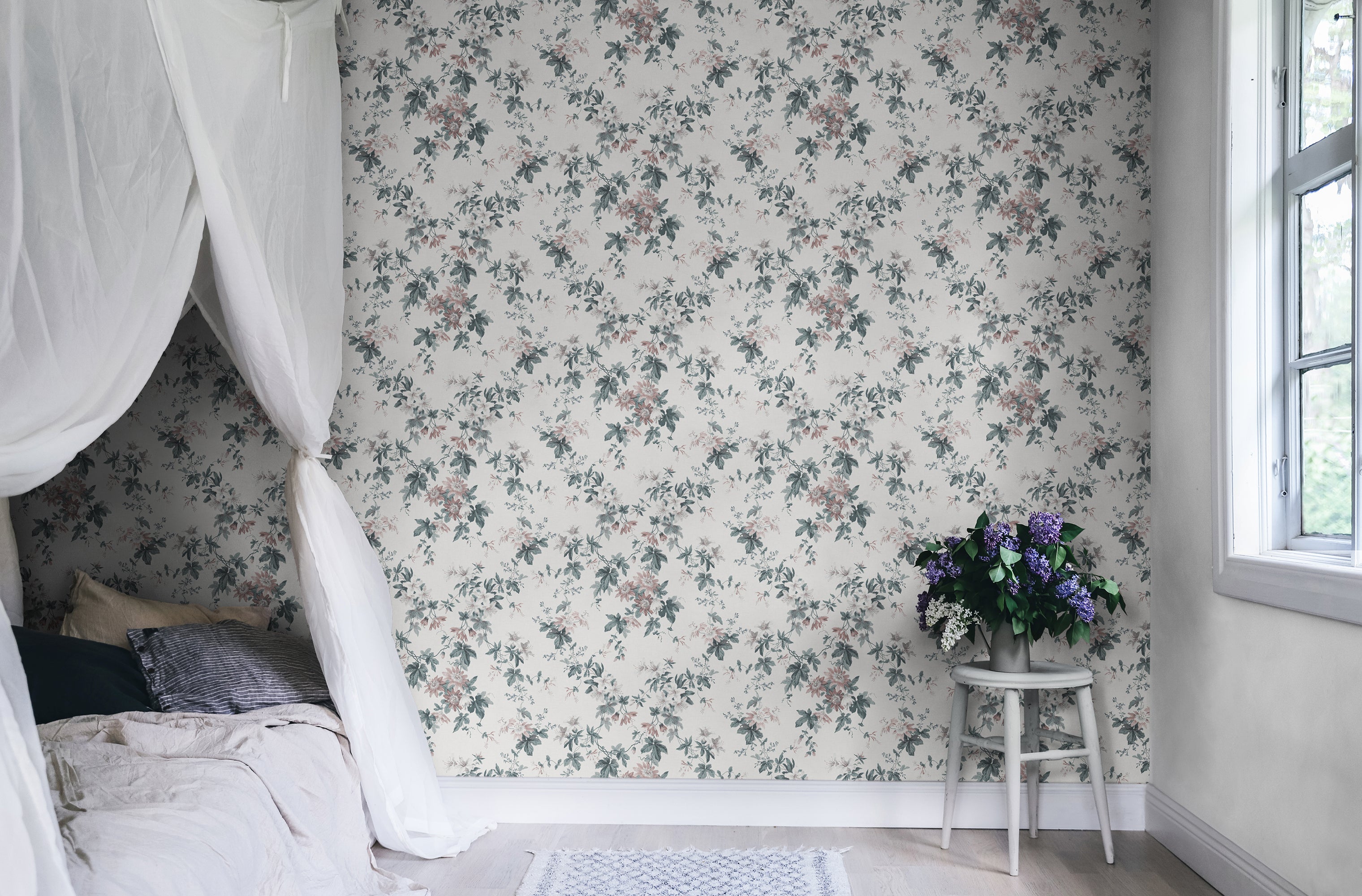 Sandra Floral wallpaper by Sandberg in cream, styled in a bedroom with the soft floral print behind a canopy bed and lilac flowers