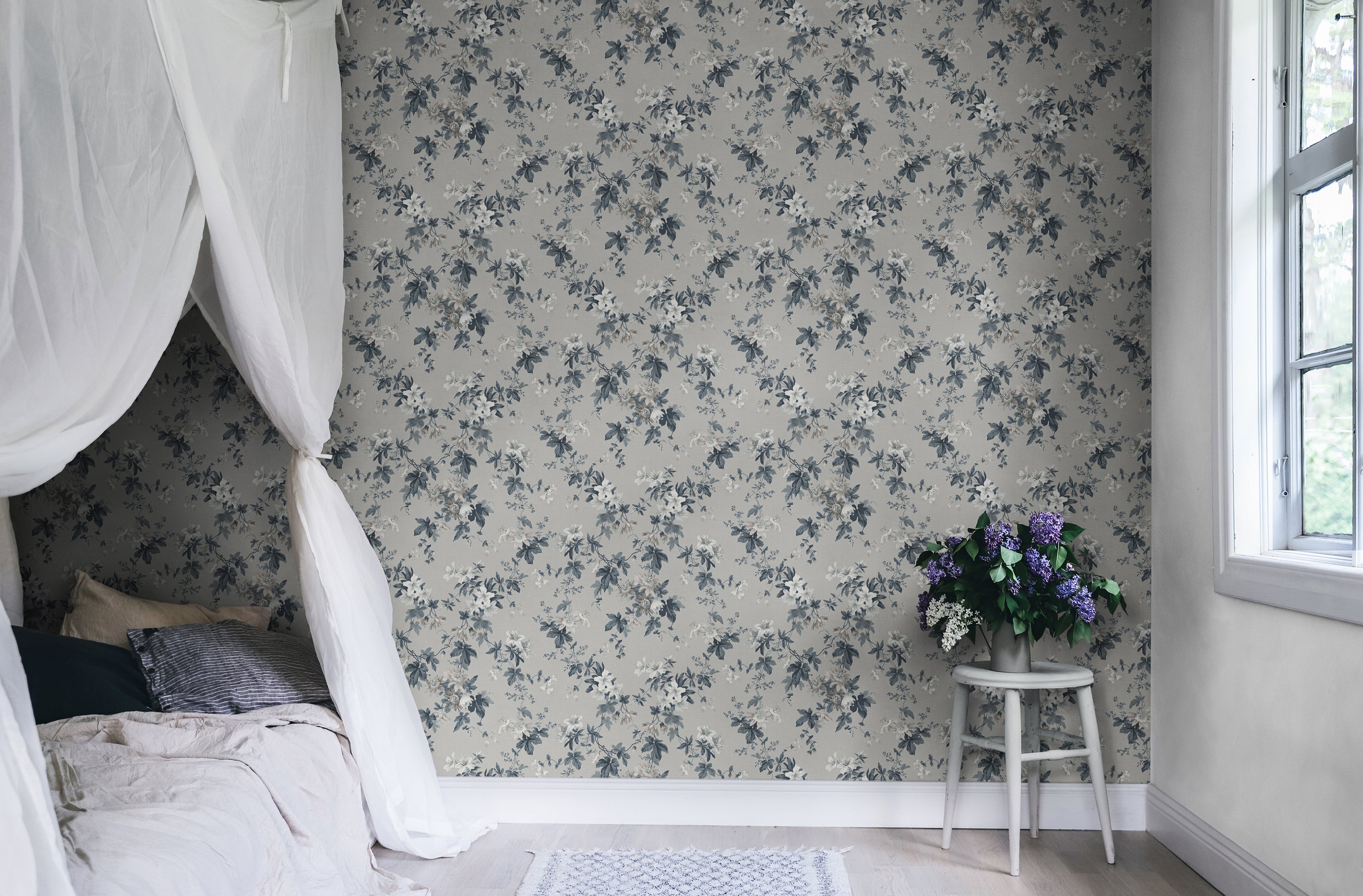 Sandra Floral wallpaper by Sandberg in soft grey, styled in a bedroom behind a white stool holding fresh lilac blooms