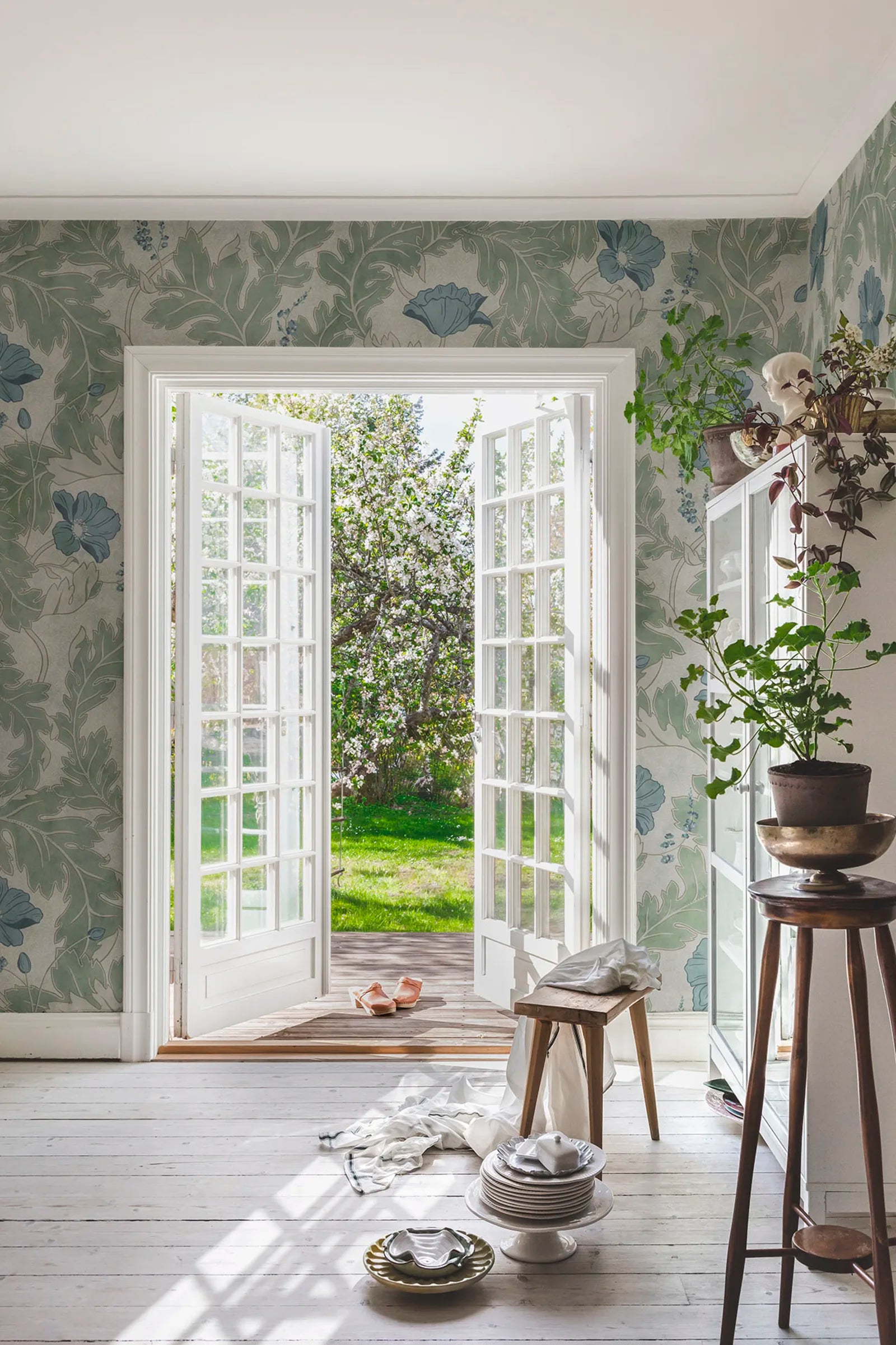 Poppy wallpaper by Sandberg in sage green with blue poppies, styled in a sunlit room with open French doors to a garden