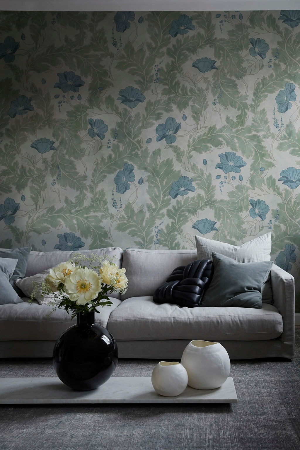 Poppy wallpaper by Sandberg in sage green with blue poppies, styled in a living room behind a grey sofa and round vases