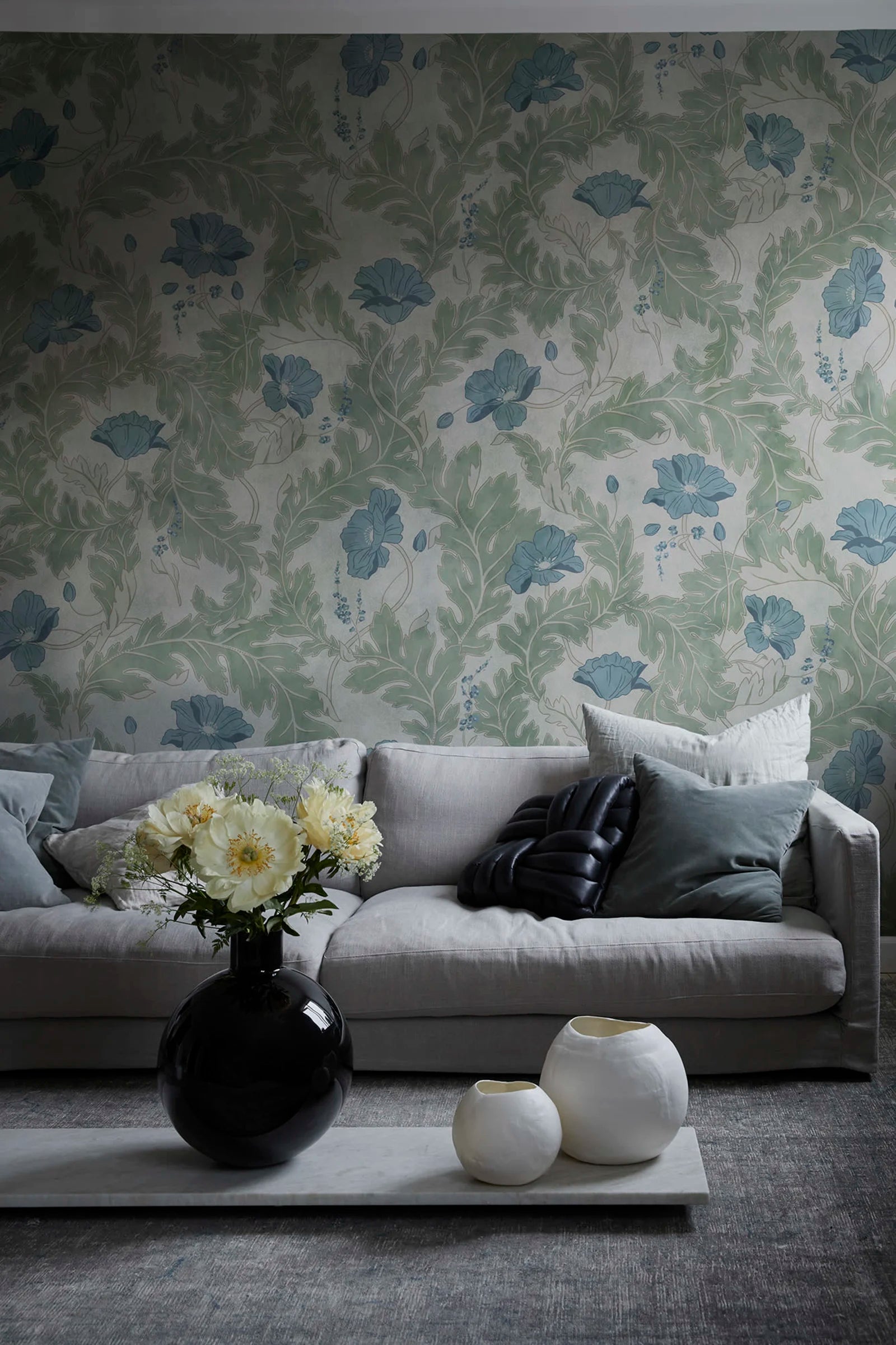 Poppy wallpaper by Sandberg in sage green with blue poppies, styled in a living room behind a grey sofa and round vases