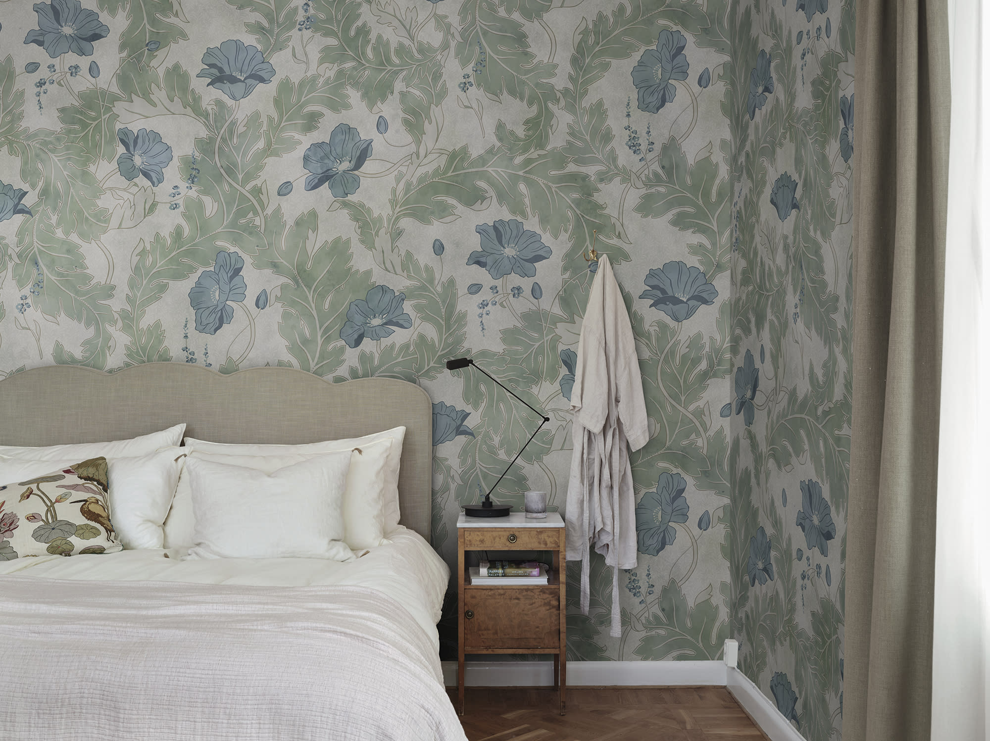 Poppy wallpaper by Sandberg in sage green with blue poppies, styled in a bedroom behind a linen headboard and timber side table