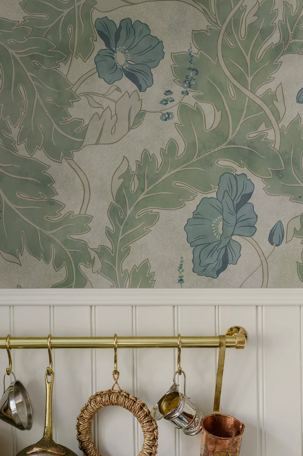 Poppy wallpaper by Sandberg in sage green with blue poppies, styled above white panelling and a brass rail of hanging kitchenware