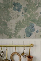 Poppy wallpaper by Sandberg in sage green with blue poppies, styled above white panelling and a brass rail of hanging kitchenware