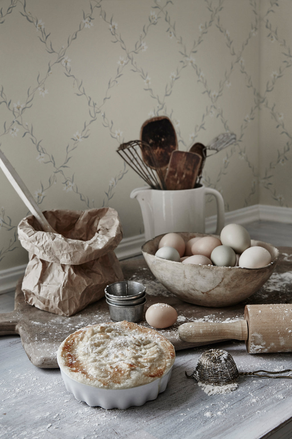 Ewa wallpaper by Sandberg in pale sage trellis floral behind a rustic baking still life with eggs and pie