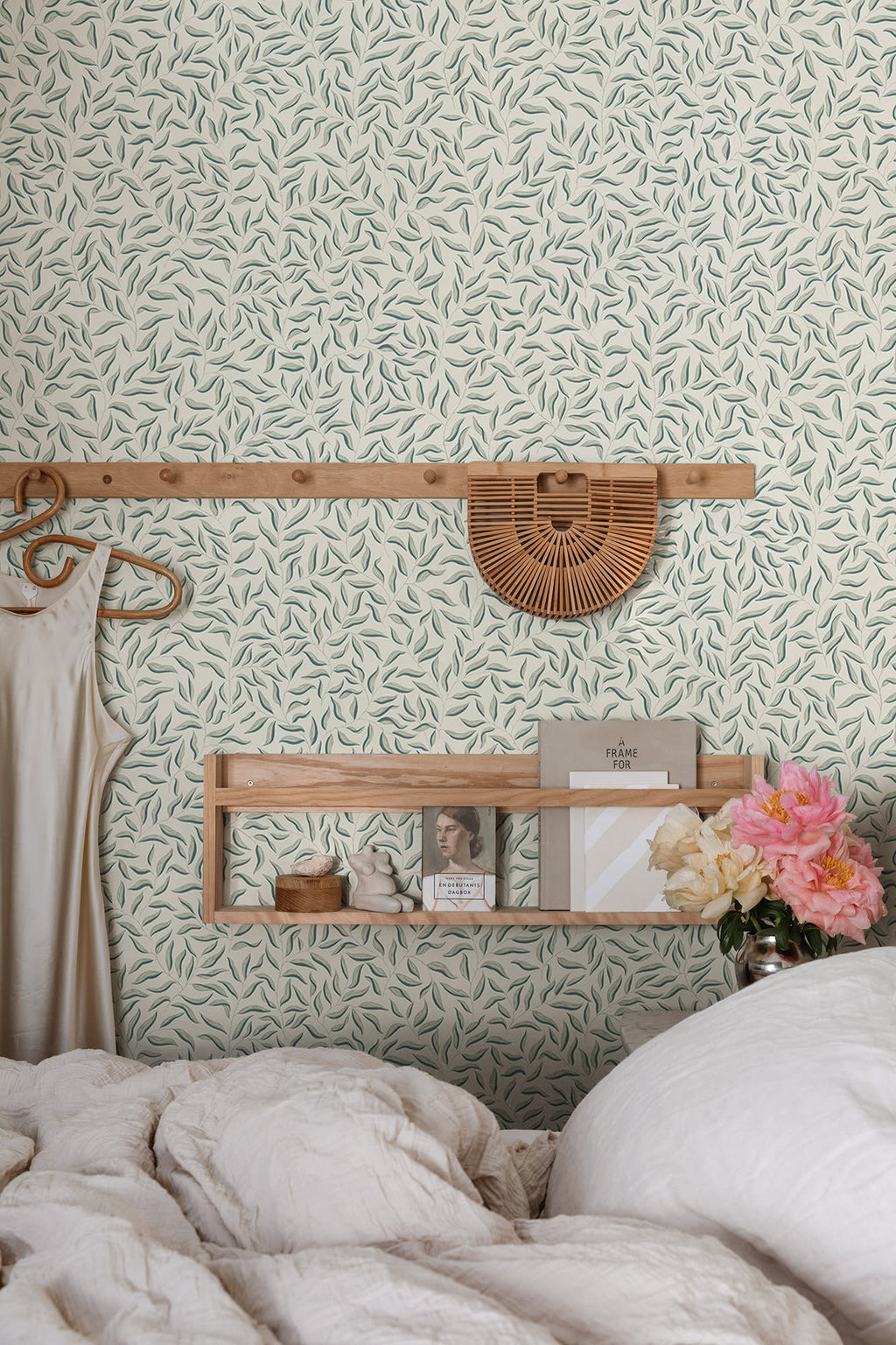Karolina sage-green leaf wallpaper by Sandberg above a bed with oak shelf and peonies in a styled bedroom