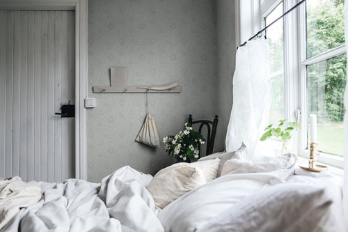 Anton wallpaper by Sandberg in pale grey behind a bed by a sunny window in an airy bedroom