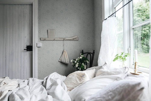 Anton wallpaper by Sandberg in soft grey behind a wall shelf and bed beside green garden views