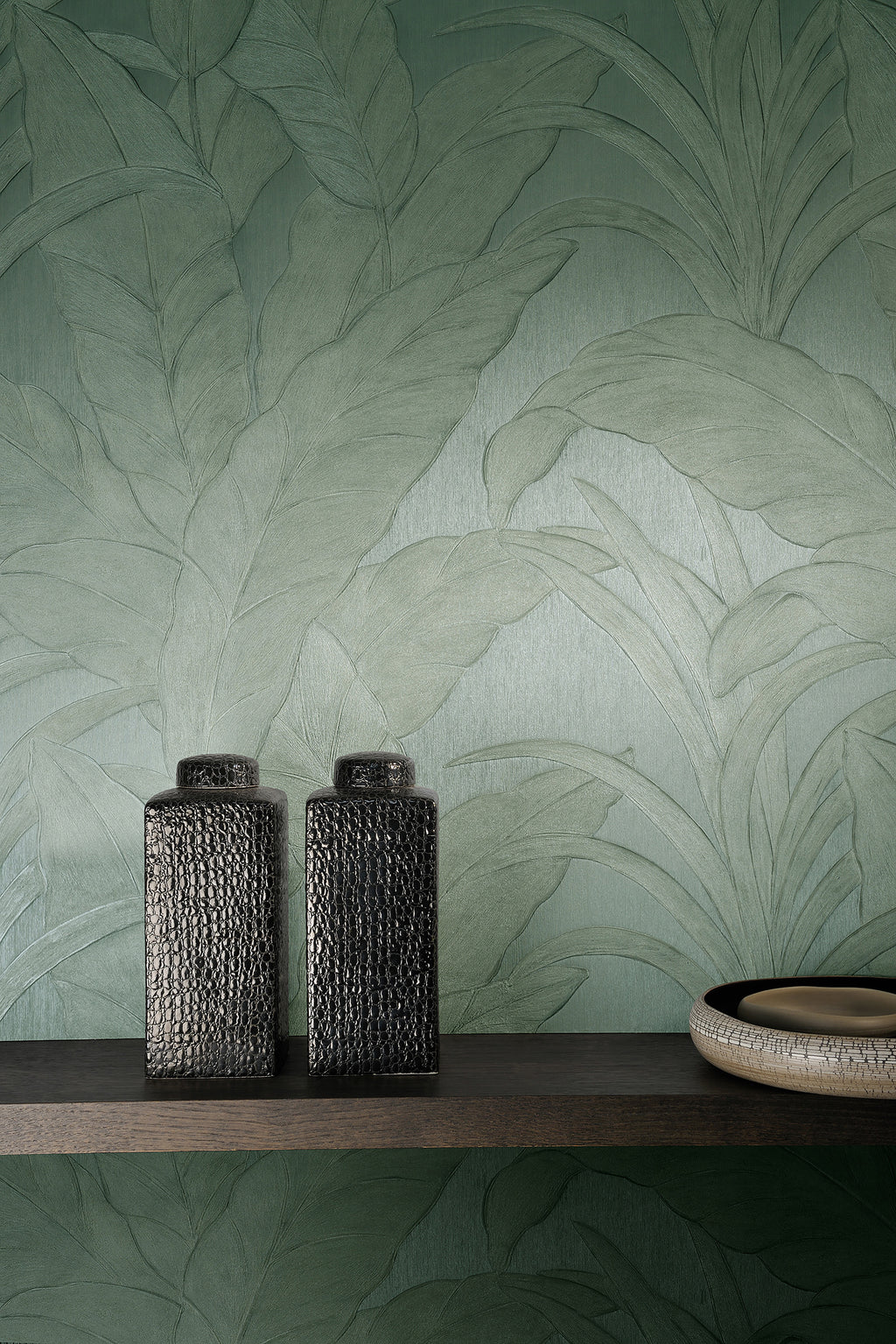 Musa wallpaper by Arte in Green behind a dark shelf holding two textured black vases and a bowl