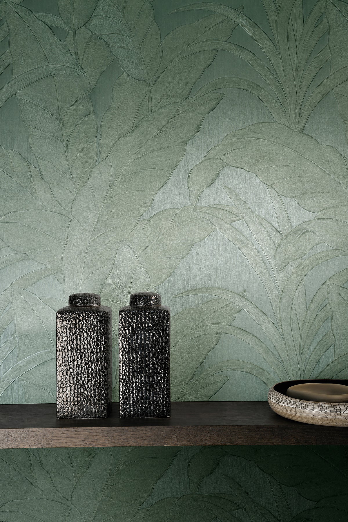 Musa wallpaper by Arte in Green behind a dark shelf holding two textured black vases and a bowl