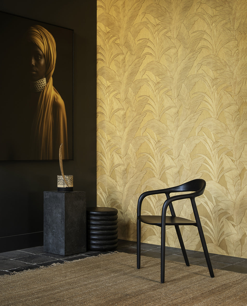 Musa wallpaper by Arte in Gold in an entry with a black chair, stone plinth and a framed portrait