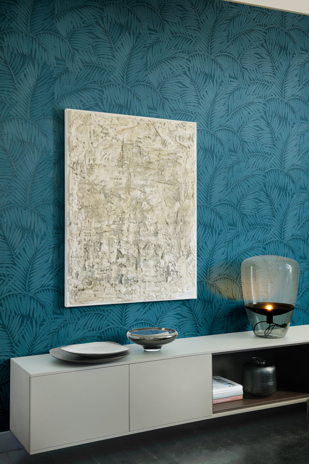 Sabal wallpaper by Arte in Teal behind a pale sideboard with a framed textured artwork and a glass lamp