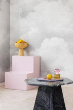 Fluffy Clouds wall mural by Rebel Walls in grey, behind a pink display plinth and marble side table with fruit