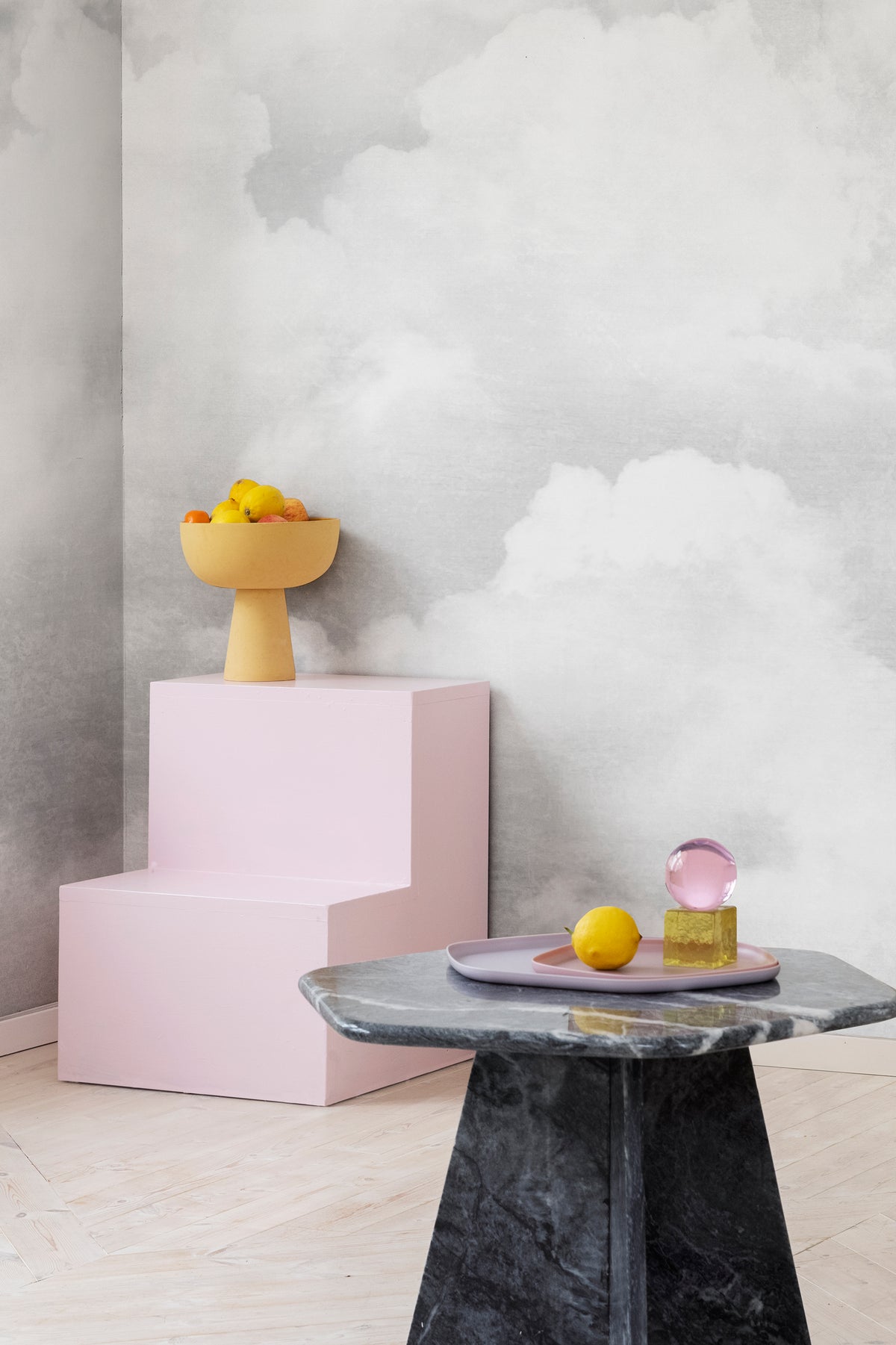 Fluffy Clouds wall mural by Rebel Walls in grey, behind a pink display plinth and marble side table with fruit