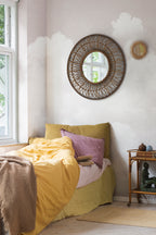 Fluffy Clouds wall mural by Rebel Walls in blush pink, behind a bed with a rattan mirror and mustard linen