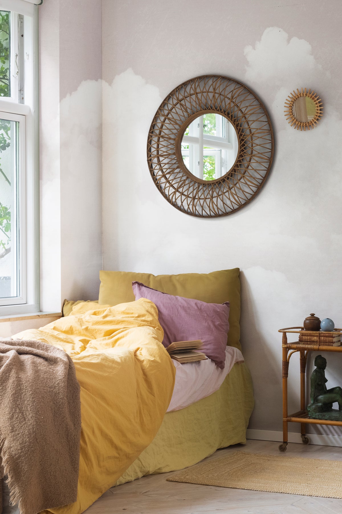 Fluffy Clouds wall mural by Rebel Walls in blush pink, behind a bed with a rattan mirror and mustard linen