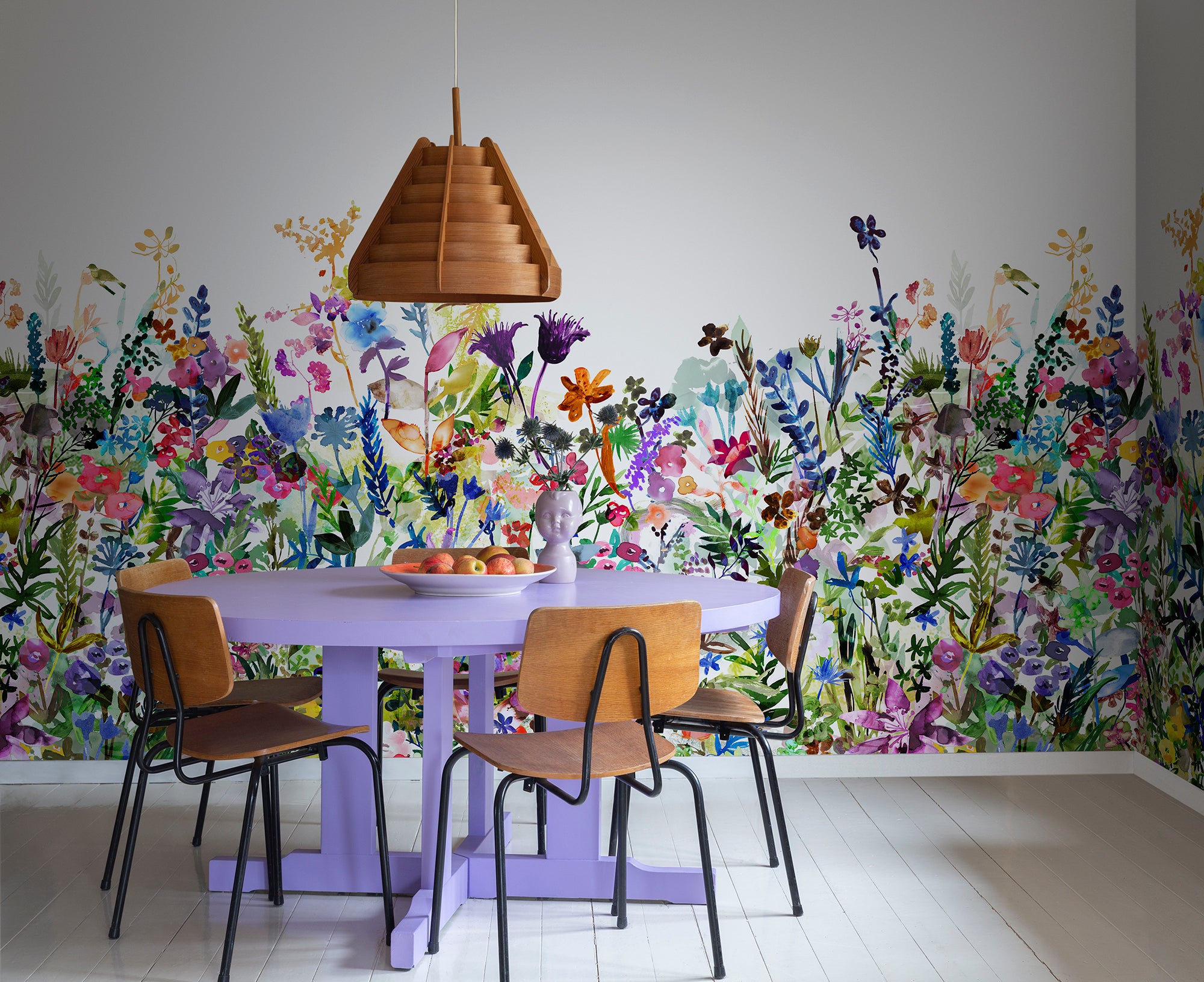May Meadow mural by Rebel Walls, rainbow wildflower meadow in a dining room behind a lilac table with timber chairs