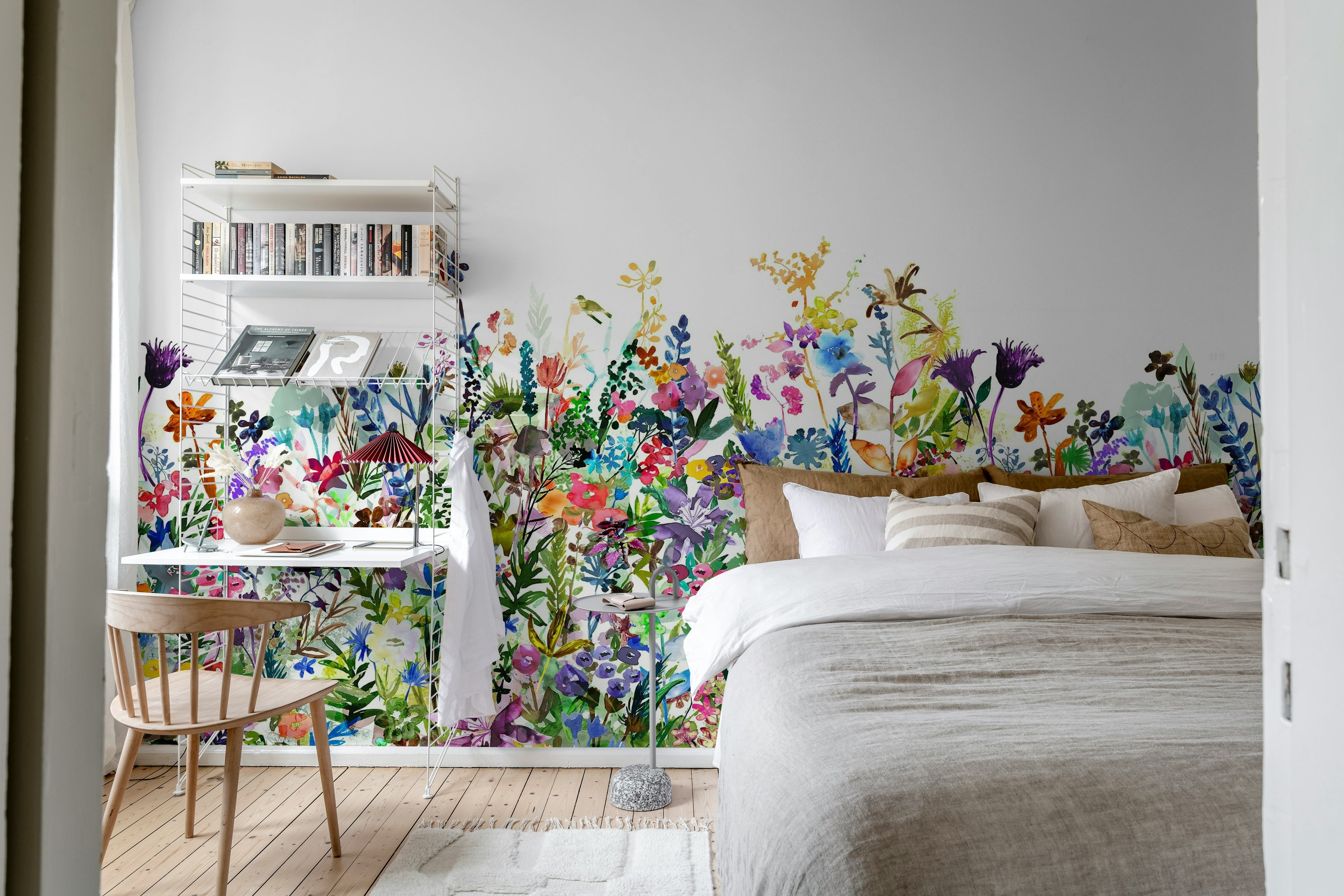 May Meadow mural by Rebel Walls, vivid wildflower meadow in a bedroom behind a linen bed with a corner desk and bookshelf