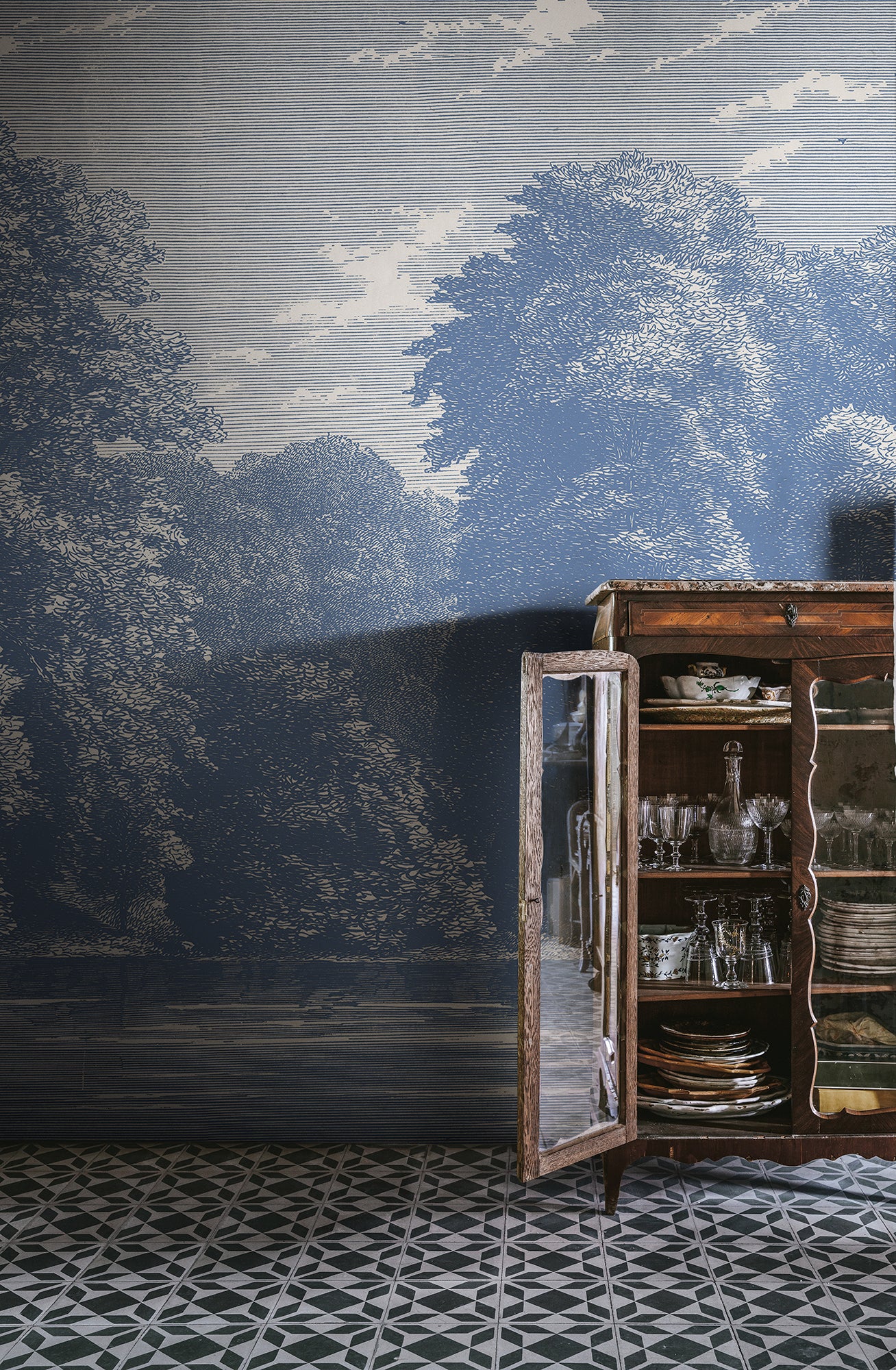 Spring Season wall mural by Rebel Walls in dusty blue, engraved woodland and clouds styled beside a glazed dresser on patterned tiles
