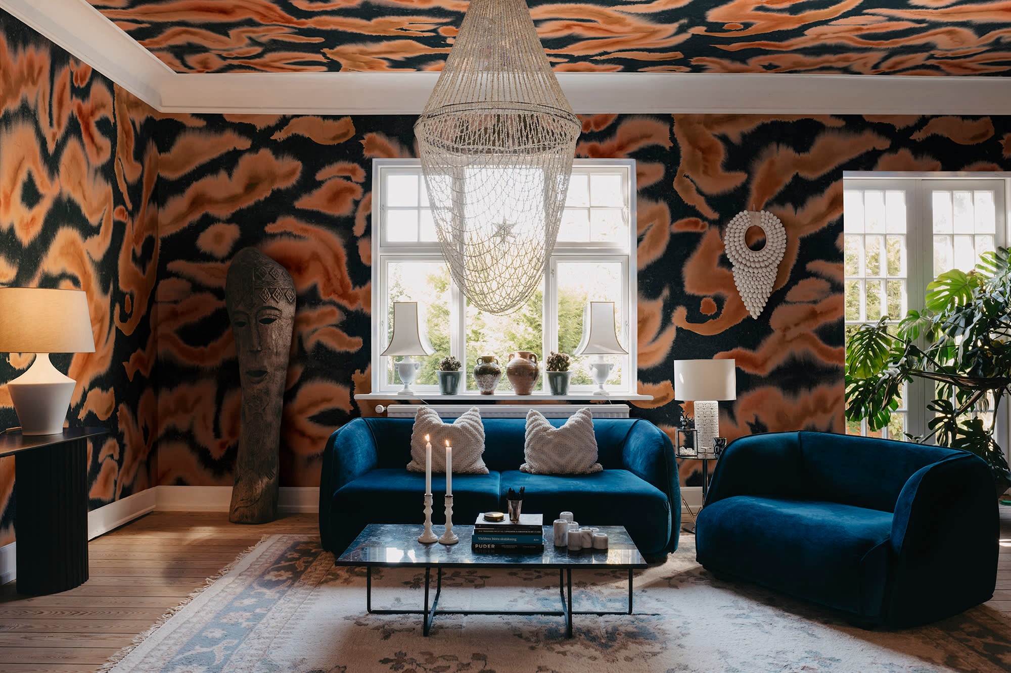 Guppy wall mural by Rebel Walls in orange and navy swirls, in a living room with blue velvet sofas and beaded chandelier