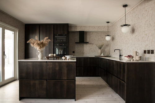 Anna wallpaper by Sandberg in beige across a moody kitchen with dark cabinetry and a sunlit doorway