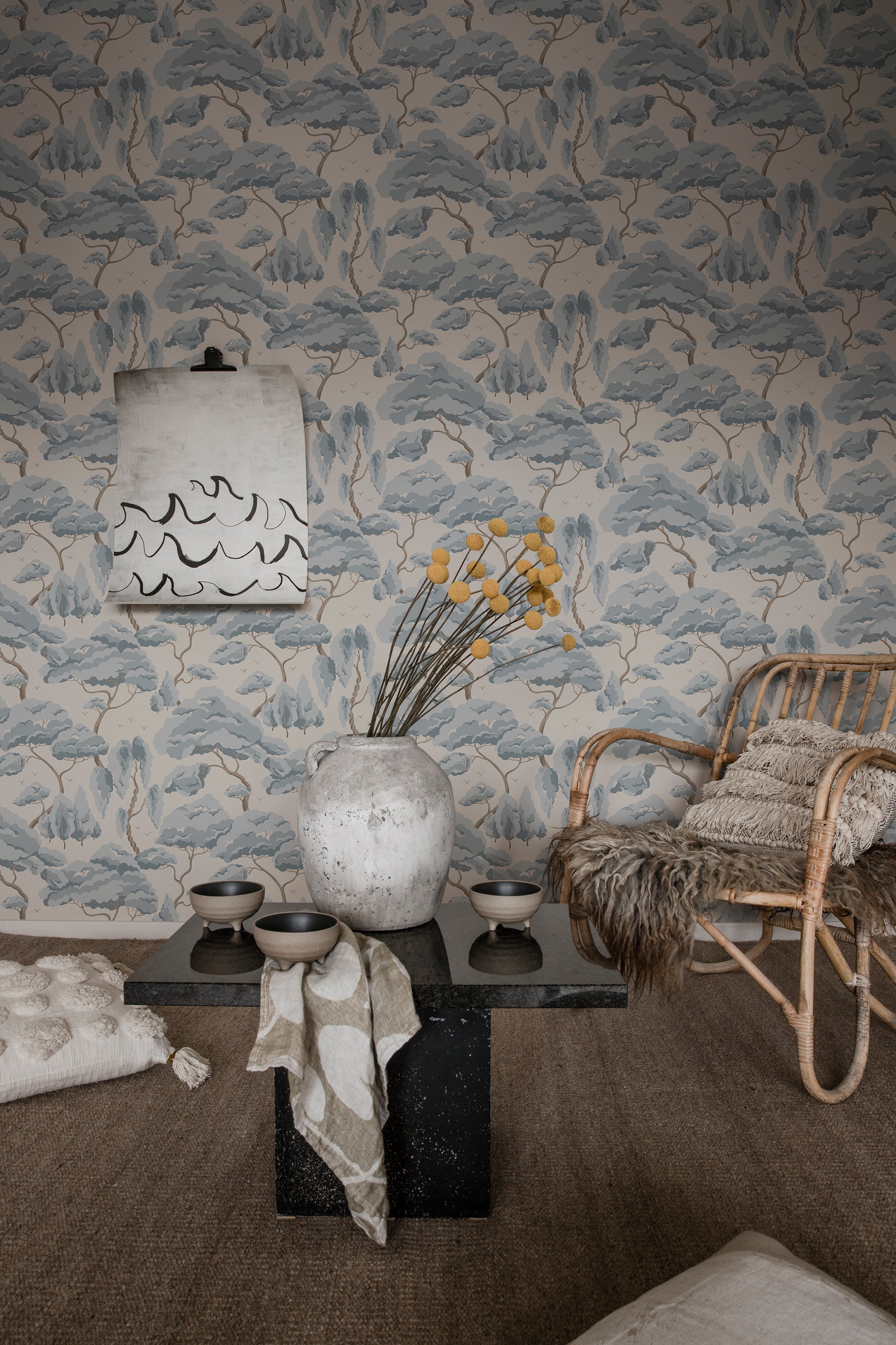 Blue-grey Kristoffer Forest tree wallpaper by Sandberg behind a black plinth table, vase and rattan armchair