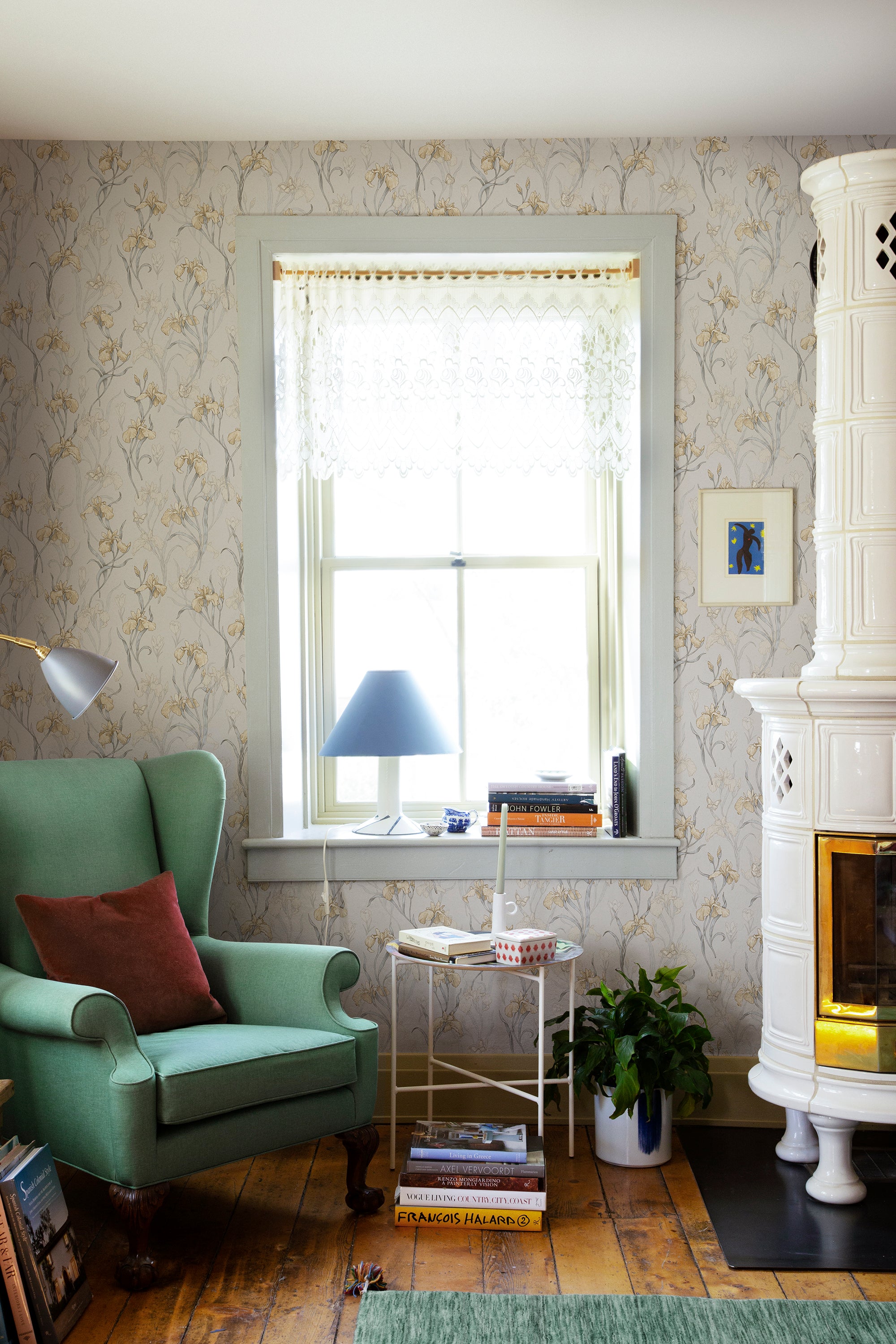 Iris wallpaper by Sandberg in soft gold styled in a cosy living room with a green wingback chair, lace-curtained window and white tiled stove