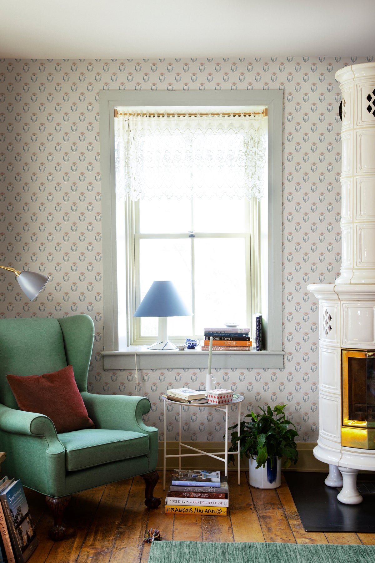 Maskrosen wallpaper by Sandberg in a cosy reading corner with a green wingback chair, tiled stove and lace-blind window