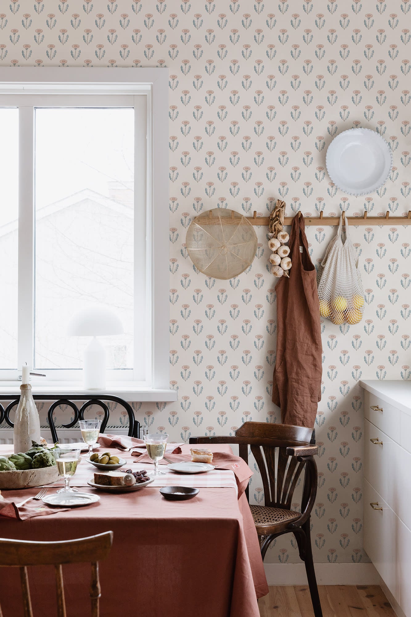 Maskrosen wallpaper by Sandberg in pink dandelion print styled in a kitchen with a rust tablecloth, peg rail and window
