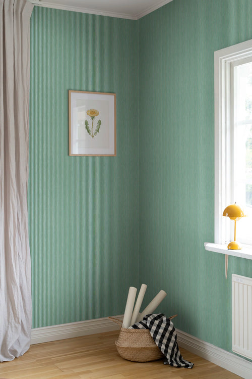 Brita Stripe wallpaper by Sandberg in green, styled in a corner with framed botanical print, basket and yellow lamp
