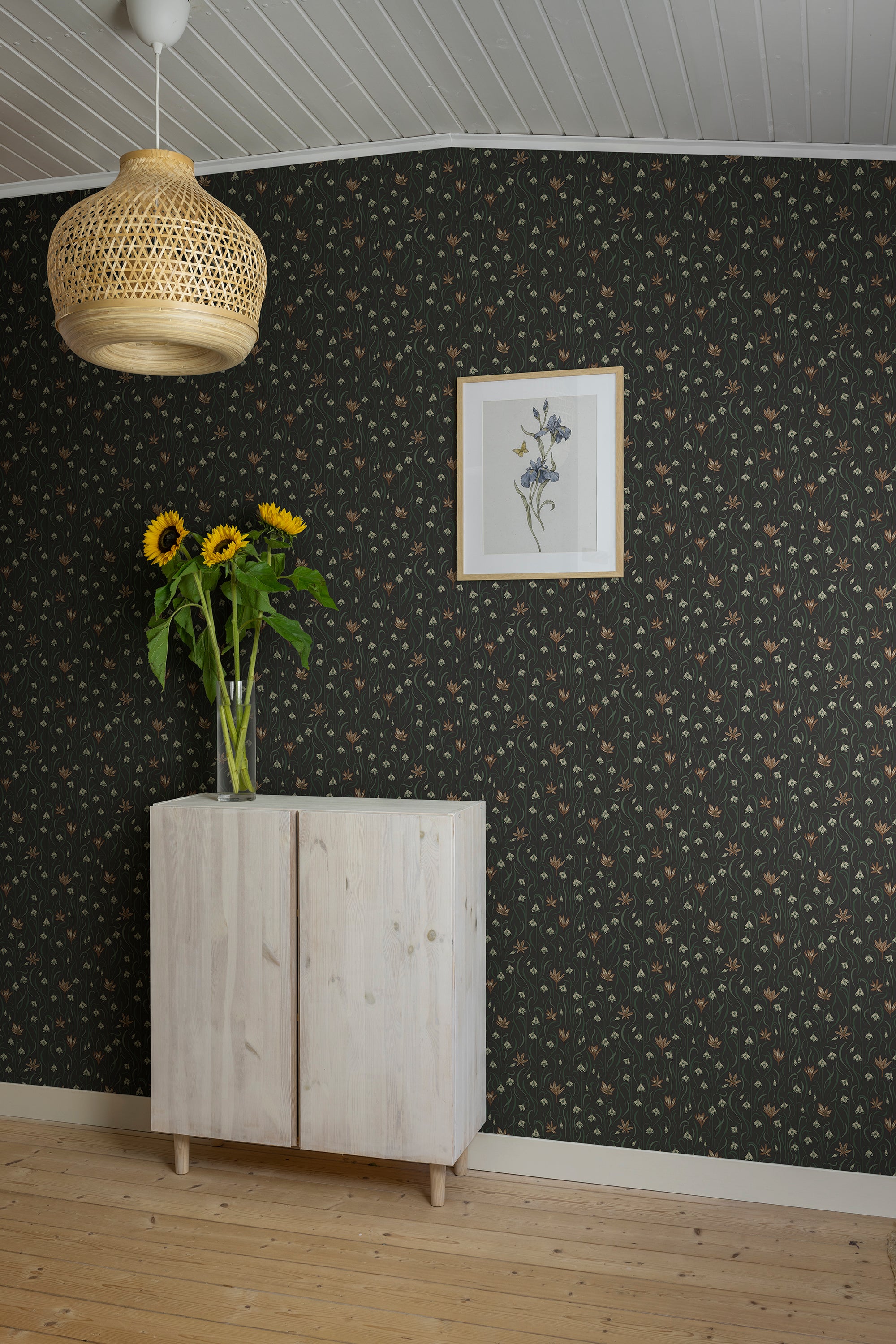 Signe wallpaper by Sandberg in charcoal behind a pale cabinet with sunflowers, a rattan pendant light and framed botanical print