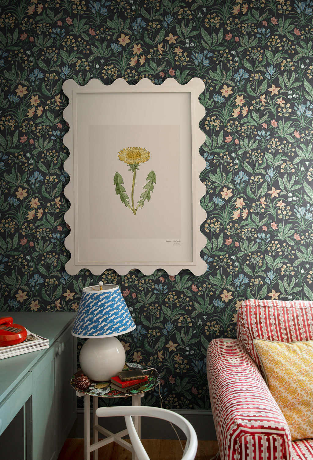 Huset i Solen wallpaper by Sandberg behind a scalloped-framed botanical print, lamp and striped sofa in a styled corner