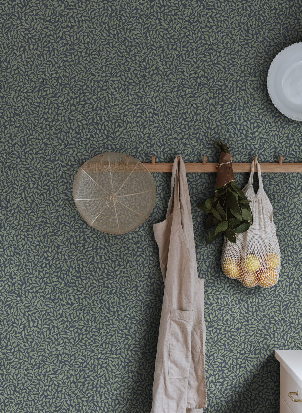 Sigfrid wallpaper by Sandberg in deep green on navy, styled in a kitchen behind a wooden peg rail with apron and produce