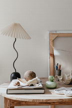 Einar wallpaper by Sandberg in greige on a wall behind a wooden table with pleated lamp and decor