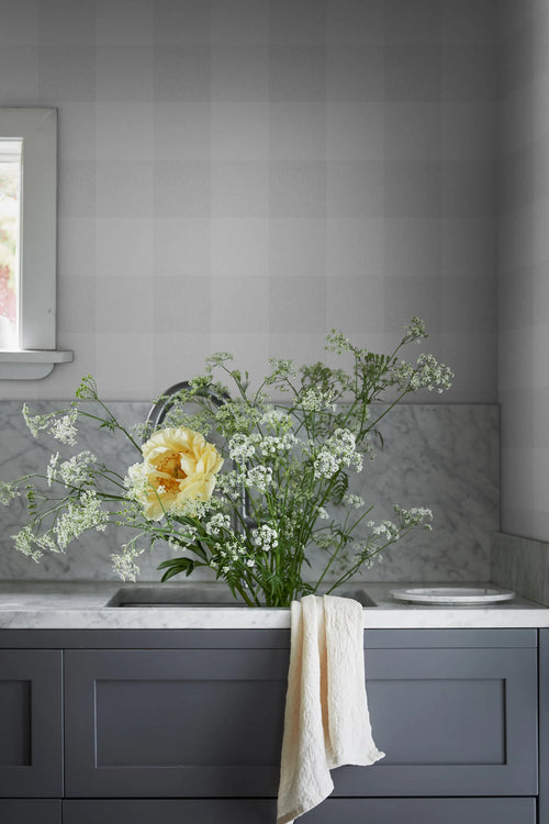 Lykke wallpaper by Sandberg in light grey buffalo check above a marble splashback and grey cabinets, yellow blooms in the