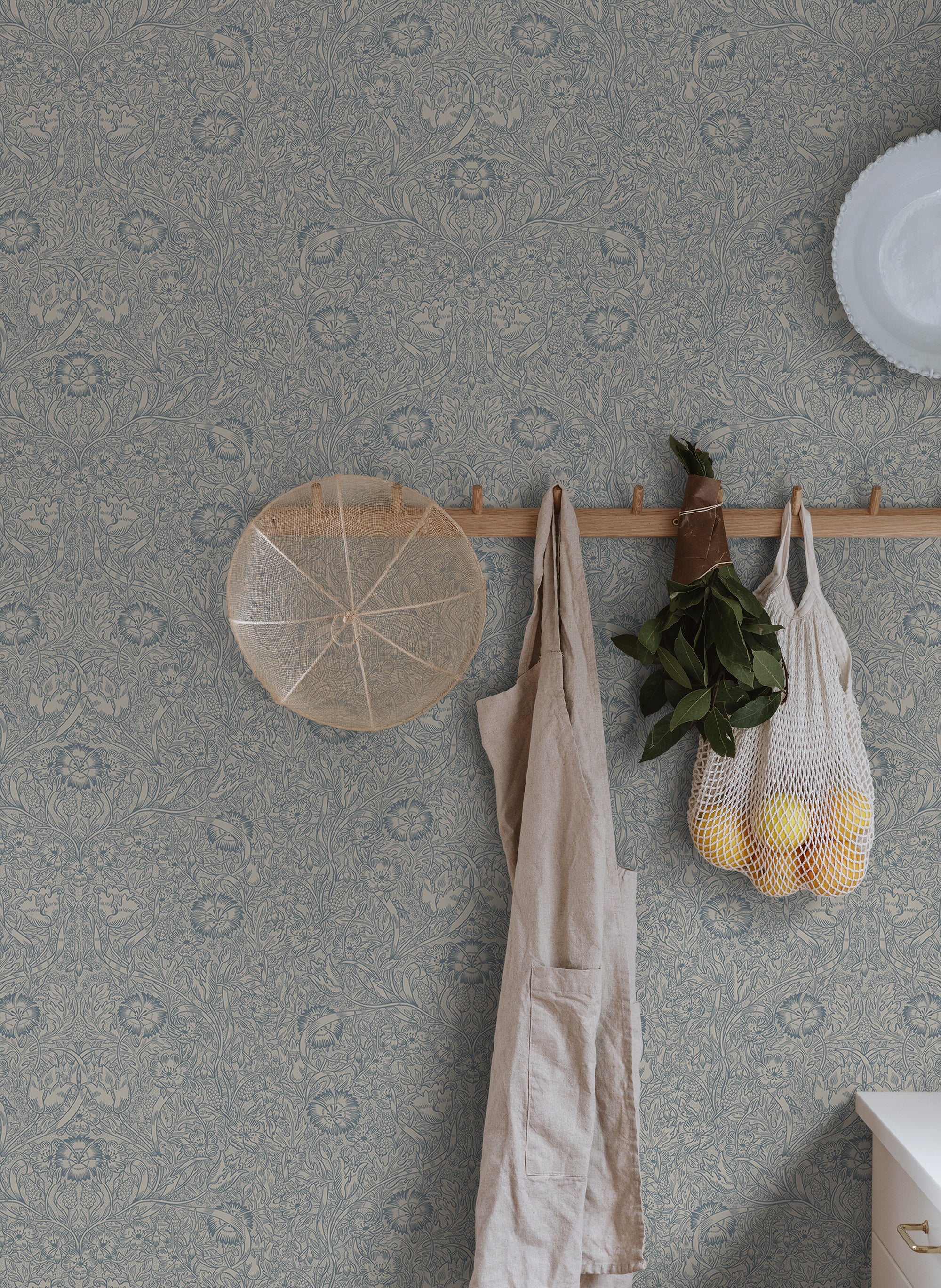 Caroline wallpaper by Sandberg in slate blue in a hallway with a peg rail holding a linen apron and string bag of lemons