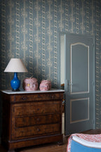 Alexandra wallpaper by Sandberg in dusty blue behind an antique wood dresser with a blue lamp and woven jars