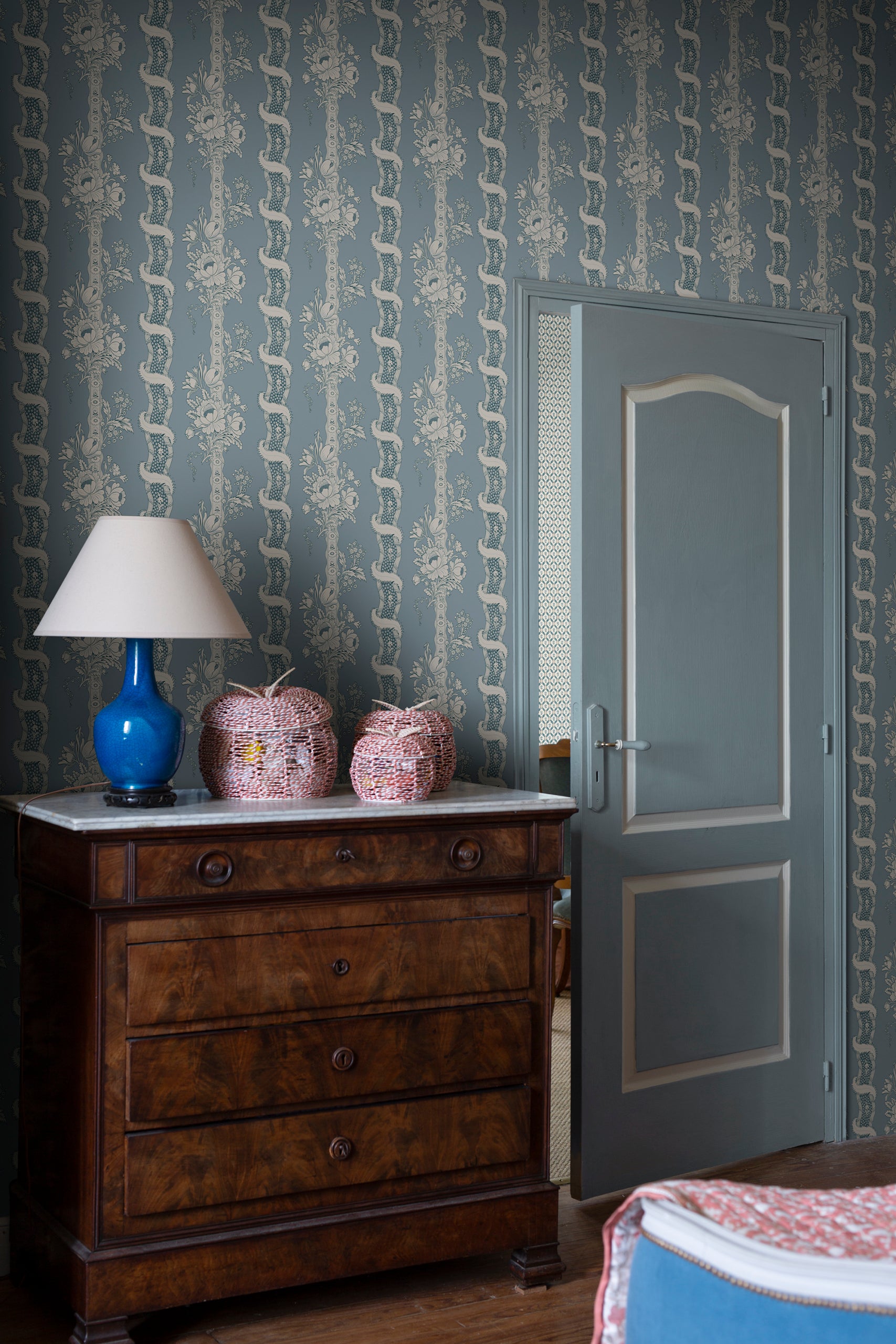 Alexandra wallpaper by Sandberg in dusty blue behind an antique wood dresser with a blue lamp and woven jars