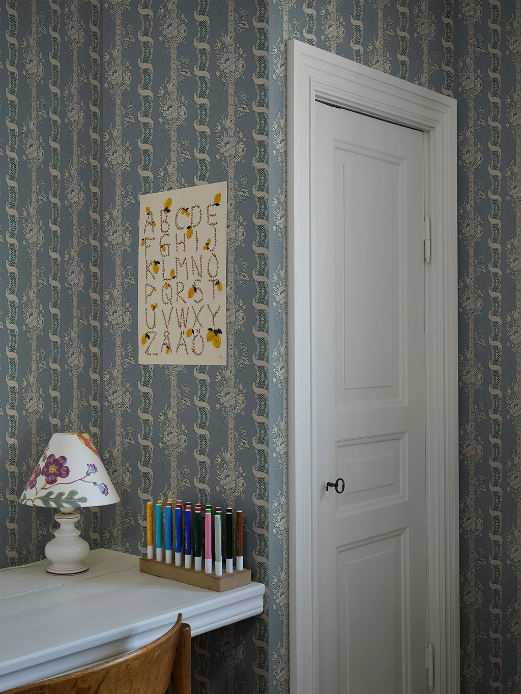 Alexandra wallpaper by Sandberg in dusty blue in a study nook with an alphabet print, lamp and pencils by a white door