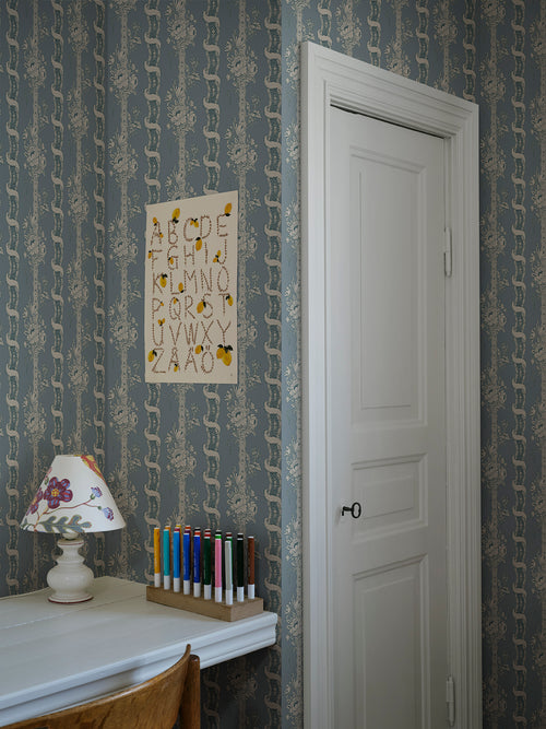 Alexandra wallpaper by Sandberg in dusty blue in a study nook with an alphabet print, lamp and pencils by a white door