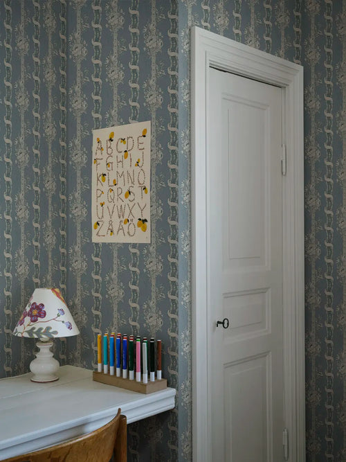 Alexandra wallpaper by Sandberg in dusty blue in a study nook with an alphabet print, lamp and pencils by a white door