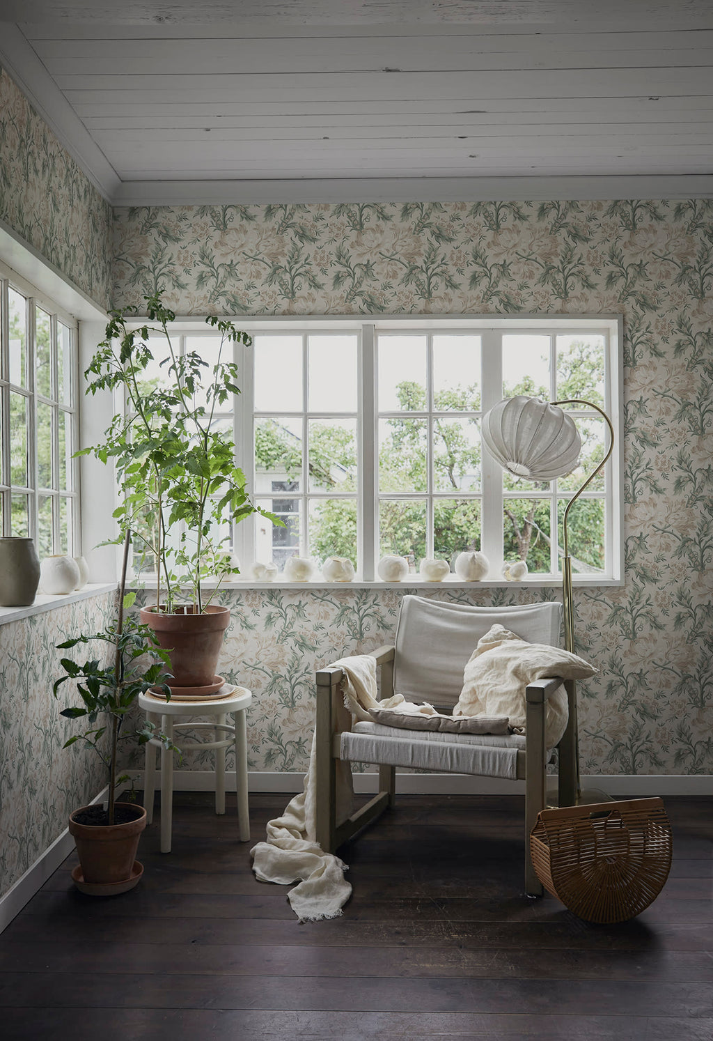 Amelia wallpaper by Sandberg lining a bright sunroom with timber floors, a linen armchair and potted plants by the window