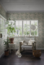 Amelia wallpaper by Sandberg lining a bright sunroom with timber floors, a linen armchair and potted plants by the window