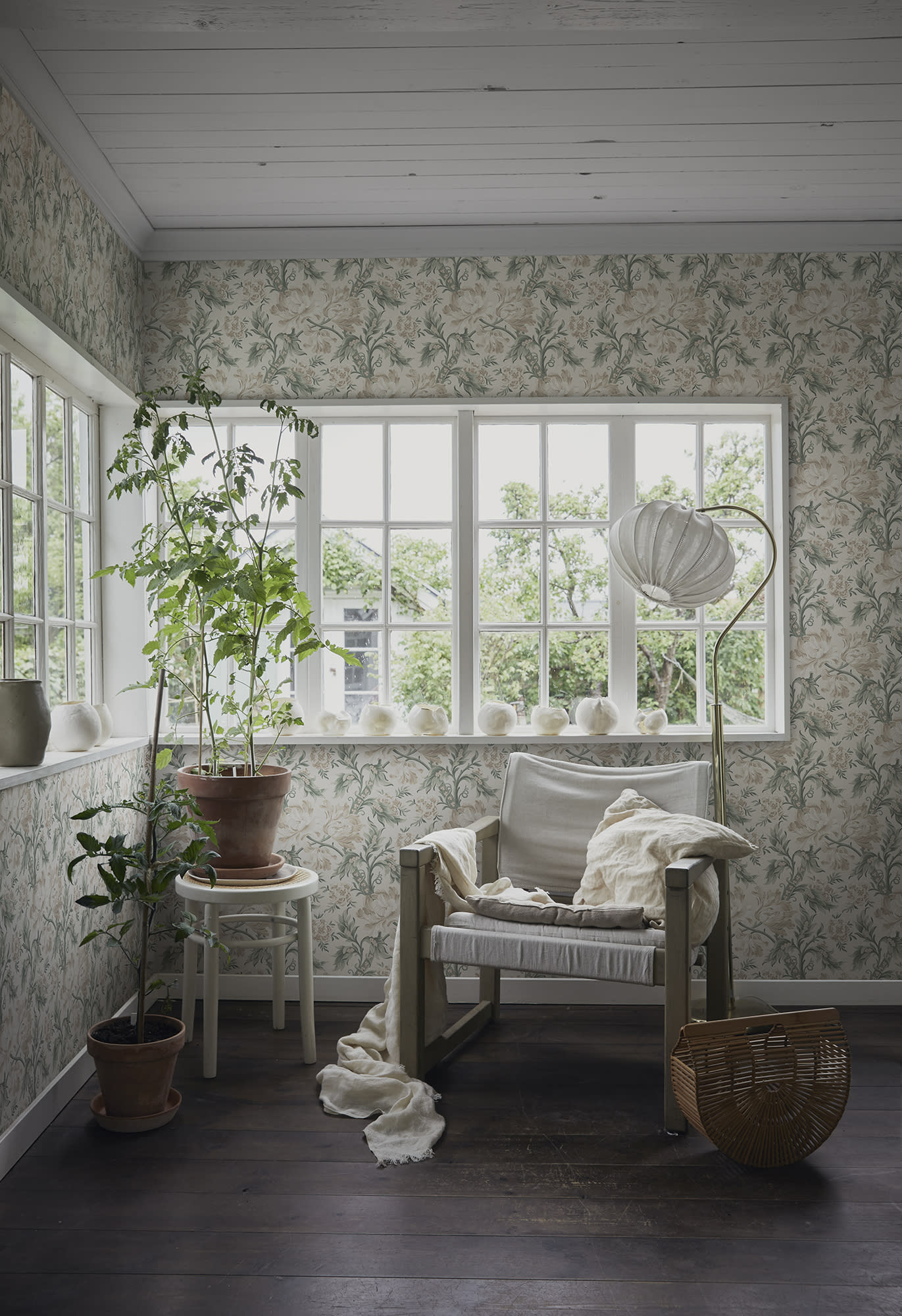 Amelia wallpaper by Sandberg lining a bright sunroom with timber floors, a linen armchair and potted plants by the window